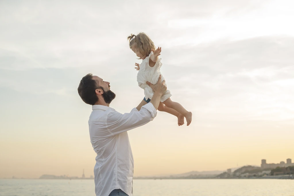 Padre levantando a su hija en sesión de fotos en la playa al atardecer.