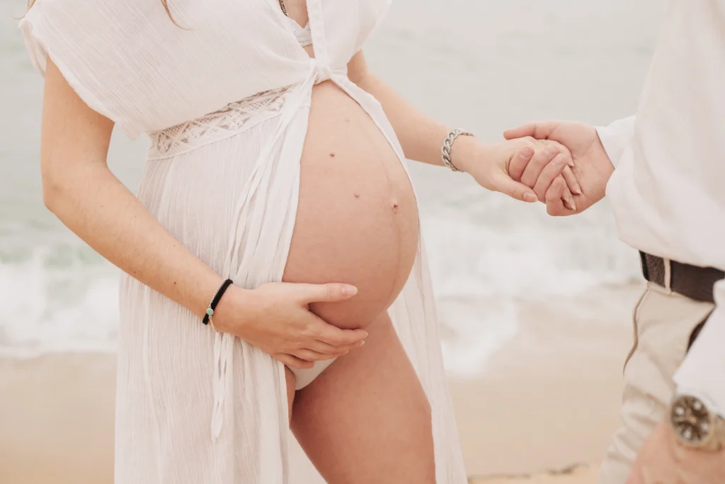 Primer plano del vientre de una mujer embarazada en la playa, con su pareja sosteniendo su mano de fondo.