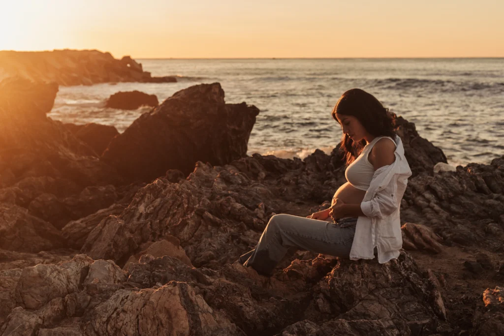 Sesión de maternidad en la playa de Barcelona al atardecer: embarazada sentada en las rocas. Sesión de fotos de embarazo en la playa de Barcelona al atardecer: mujer embarazada sentada en rocas mirando su barriga.