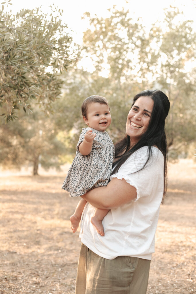 Sesión familiar en exteriores en Barcelona: madre con su bebé en brazos, sonriendo entre árboles al atardecer
