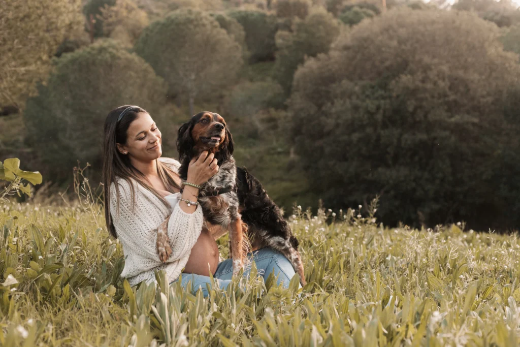 Sesión de fotos de embarazo con perro en la naturaleza: reportaje familiar natural y con mascotas. Mujer embarazada sentada en la hierba del campo sonriendo mientras acaricia a su perro
