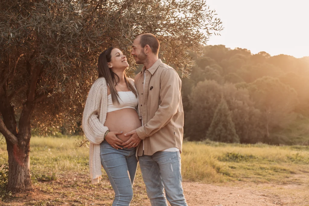 Sesión de fotos de embarazo en pareja en exteriores: conexión natural bajo la luz dorada del sol. Pareja de pie posando bajo un olivo en sesión natural de embarazo al atardecer en el campo