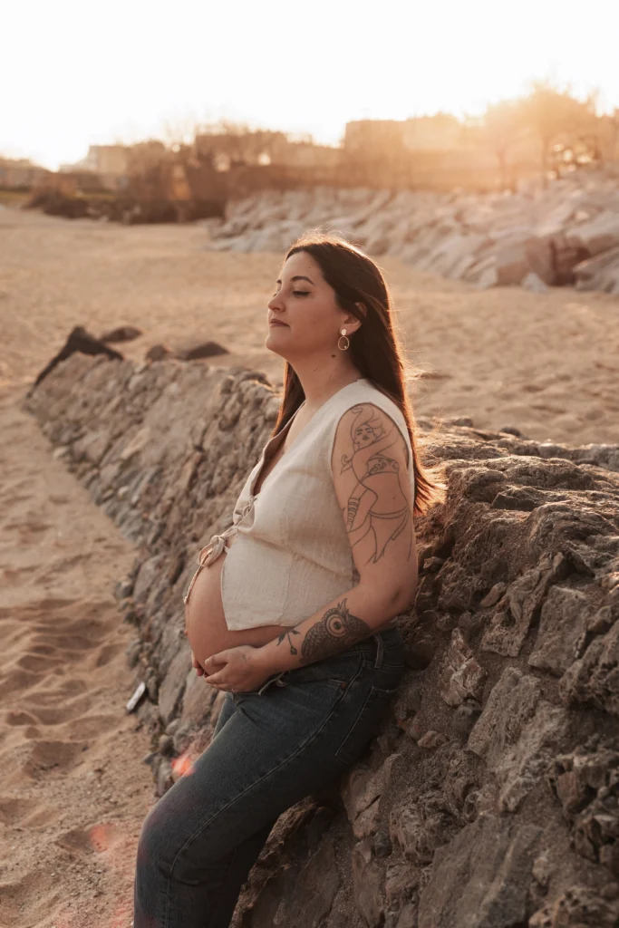 Sesión de fotos de embarazo en exterior: luz natural y estilo natural en la costa de Barcelona. Mujer embarazada con tatuajes posando apoyada en un muro de piedra en la playa al atardecer en Barcelona