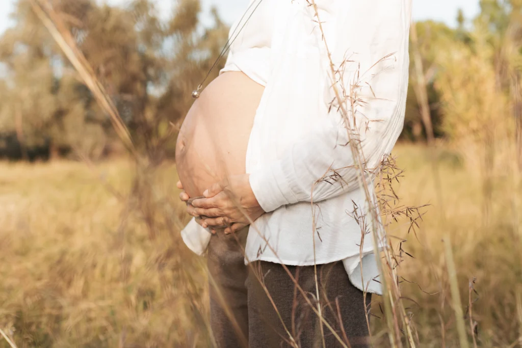 Fotografía natural de maternidad en el campo: embarazada con camisa blanca y estilo rústico. Plano detalle de barriga de embarazada en exteriores de naturaleza al atardecer en Barcelona.