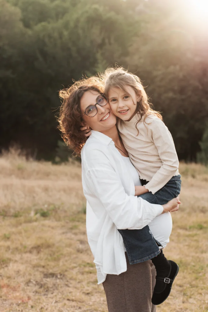 Fotos de embarazo con hija mayor en la montaña: reportaje familiar de maternidad en la naturaleza. Madre embarazada posando sonriente en el campo mientras sostiene a su hija pequeña en brazos.