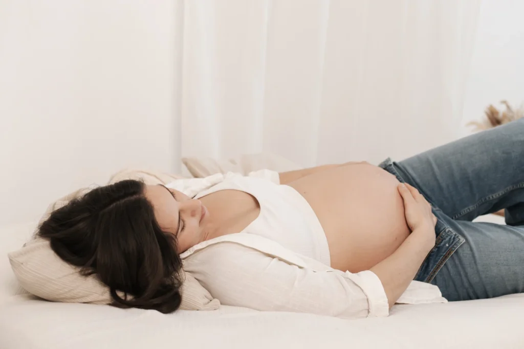 Sesión de fotos de embarazo lifestyle en estudio: pose cómoda y casual con vaqueros en Barcelona. Embarazada acostada en la cama durante sesión de fotos en estudio Barcelona.