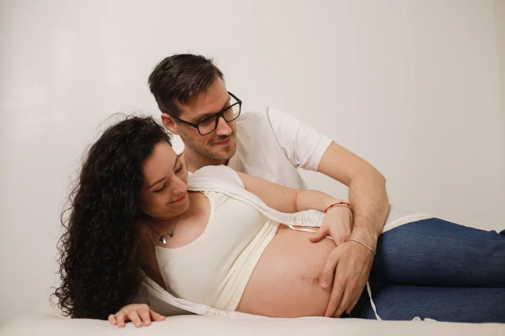 Sesión de fotos de embarazo en pareja en estudio: conexión y amor en espera de tu bebé. Futuros padres en la cama durante sesión de fotos de embarazo en estudio Barcelona.