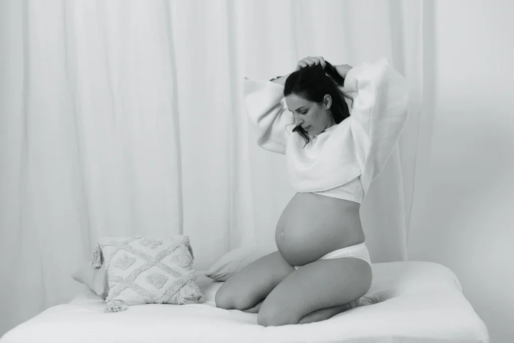 Retrato artístico de embarazada en blanco y negro: poses elegantes en estudio de maternidad. Reportaje de fotos de embarazo en estudio Barcelona, blanco y negro: mujer sentada en cama con estilo natural y luz suave.