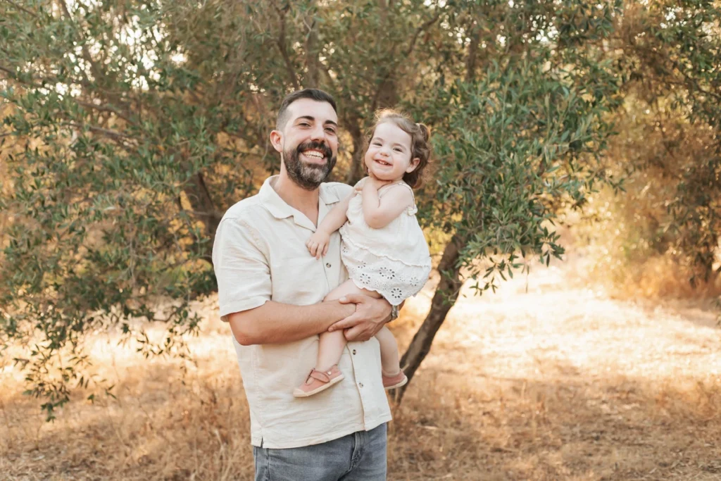Sesión de fotos familiar en el campo de Barcelona: padre sonriente sosteniendo a su hija pequeña de la mano.