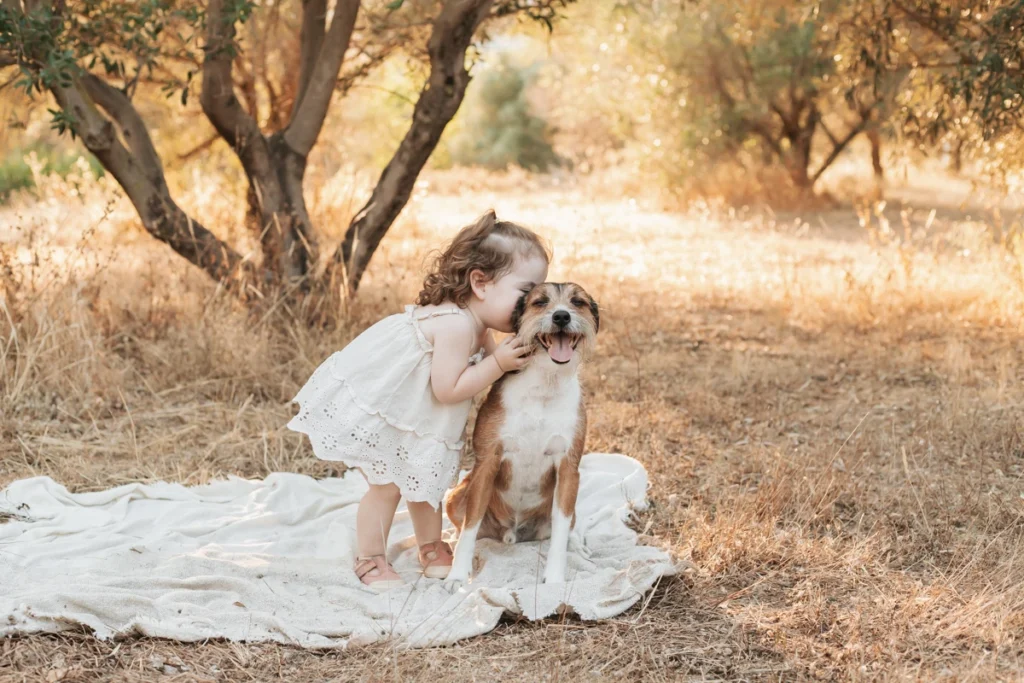 Sesión de fotos infantil natural en Barcelona: niña pequeña con vestido blanco abrazando a un perro en una manta en el campo.