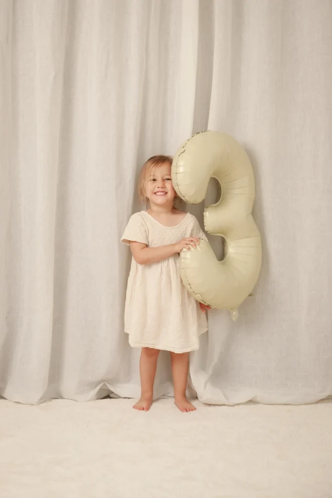Reportaje fotográfico infantil de 3 años estilo natural en Santa Coloma de Gramenet, Barcelona - Saray Ruiz Fotografía Sesión de fotos de cumpleaños para niña de 3 años sonriendo con globo gigante del número tres en estudio de Barcelona.