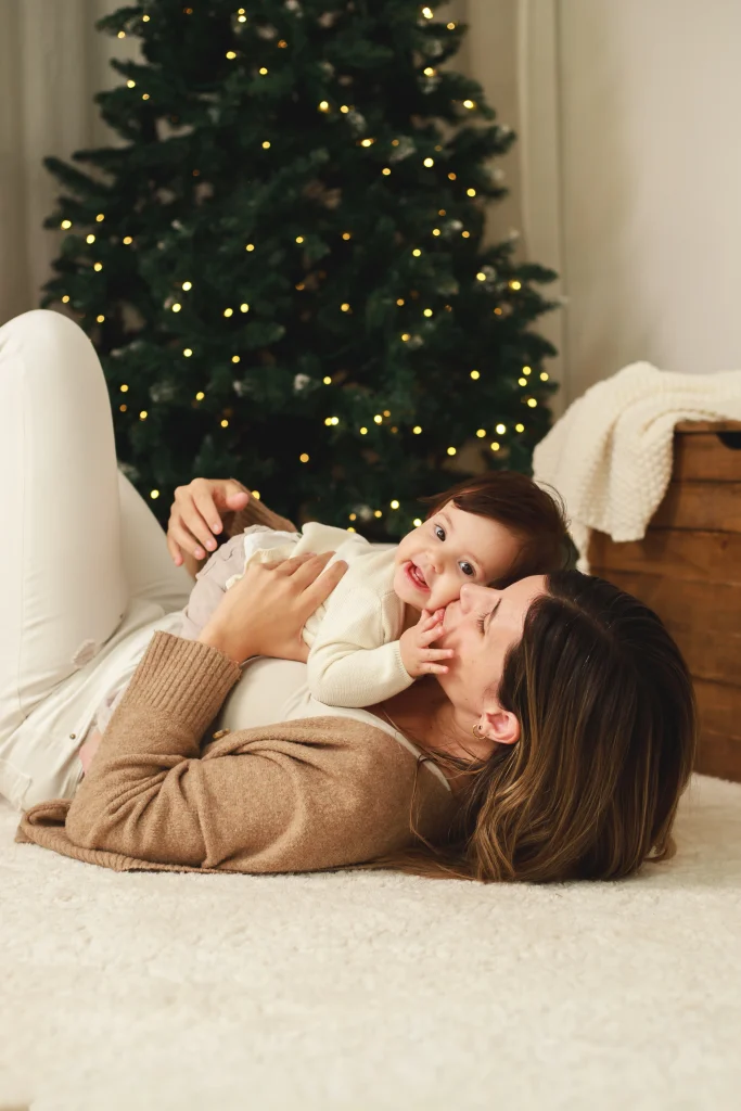 Madre tumbada en el suelo jugando y riendo con su bebé frente al árbol de Navidad decorado