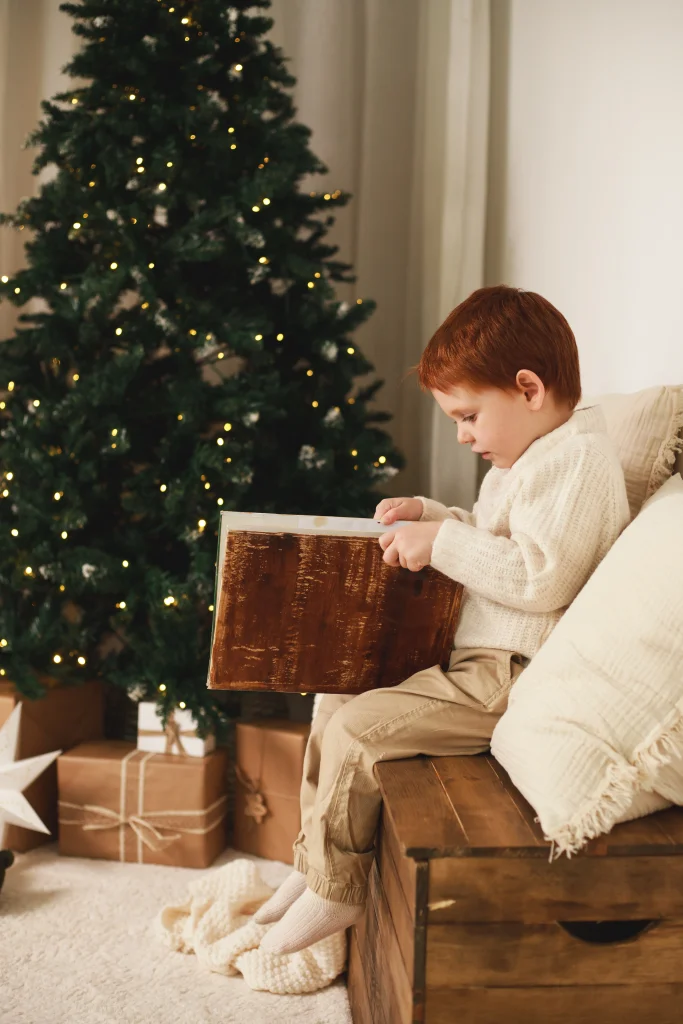 Niño sentado sobre un baúl de madera abriendo un libro junto al árbol de Navidad