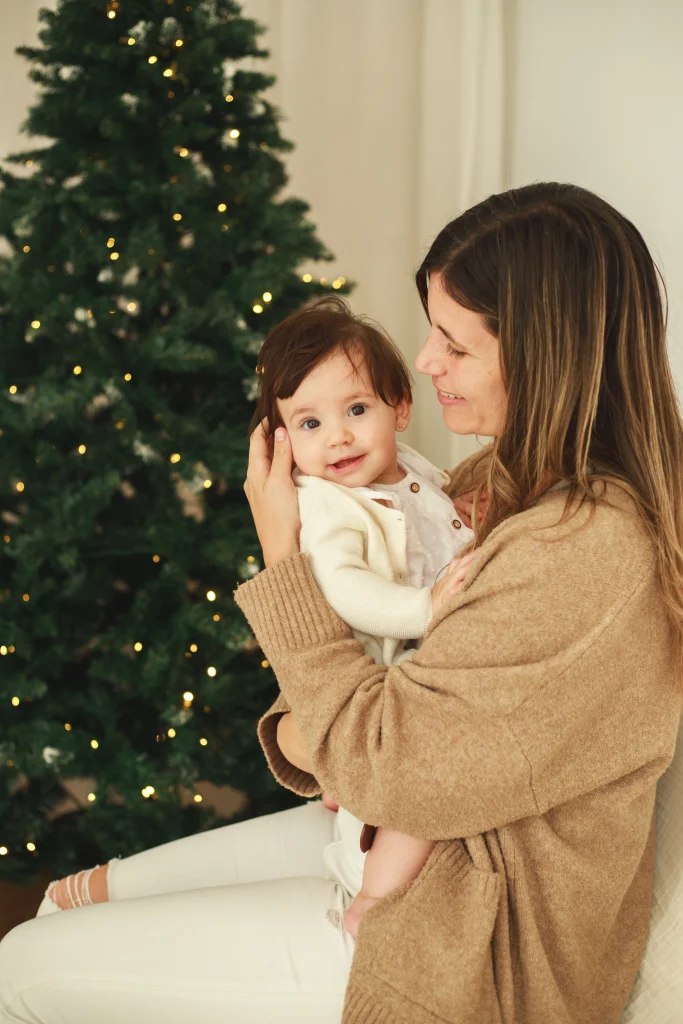 Madre sosteniendo en brazos a su bebé y mirándolo con ternura frente al árbol de Navidad