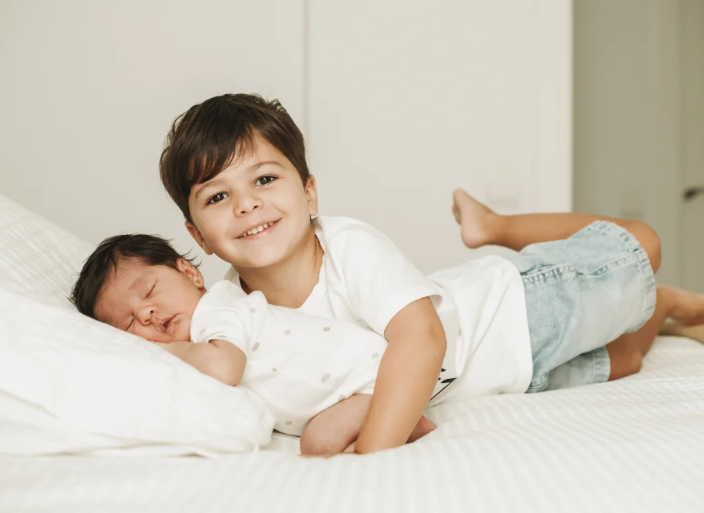 Sesión de recién nacido con hermano mayor: niño sonriente tumbado junto al bebé dormido