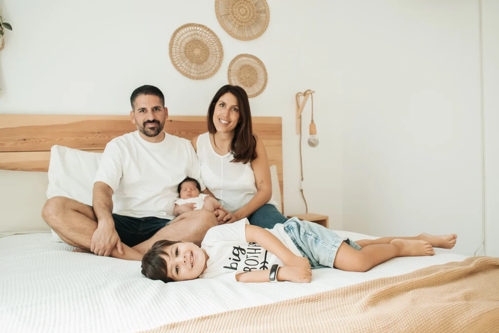 Fotografía familiar de recién nacido a domicilio en Barcelona: padres, hermano mayor y bebé en la cama